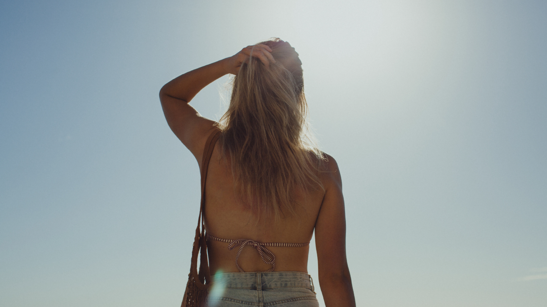 A blonde women stands facing the sun, back to camera. Her hand is in her hair and the sun and blue sky in front of her. 