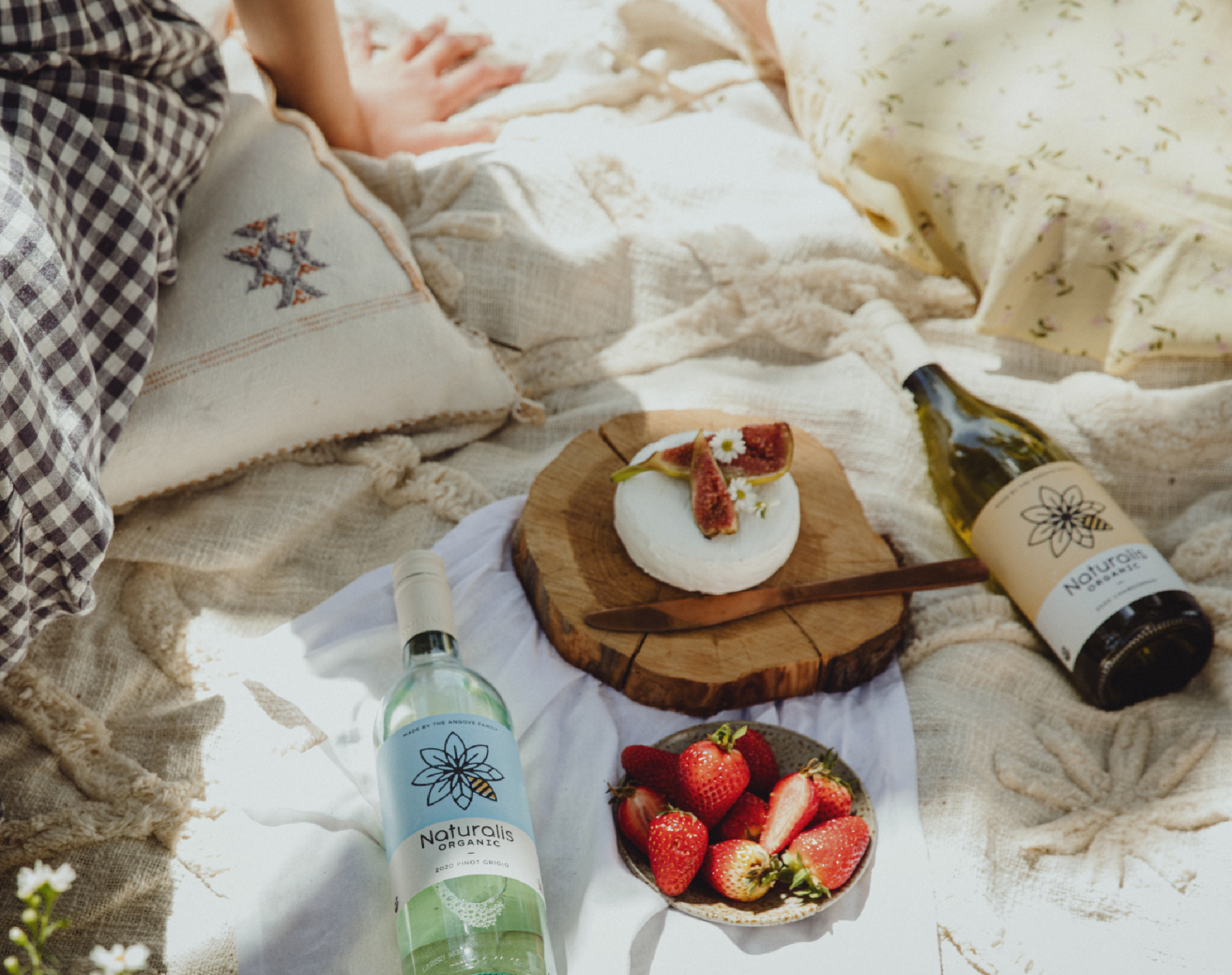 Picnic setting showing Angove's Naturalis Organic wine bottles on a soft cream linen blanket paired with soft cheese on a wood platter and strawberries in a bowl. 