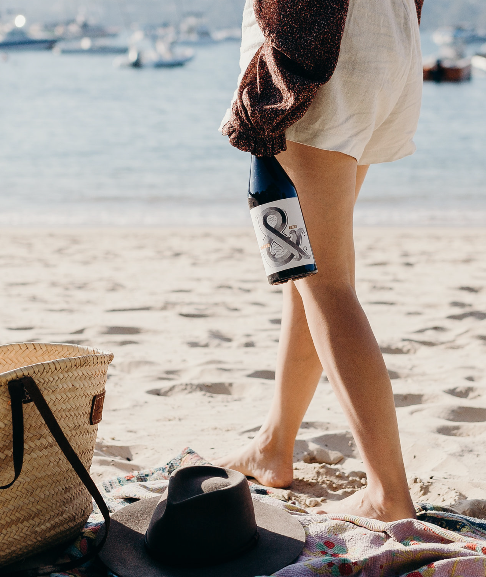 Woman walking along the beach in a pair of shorts with a bottle of Hither & Yon wine in one hand.