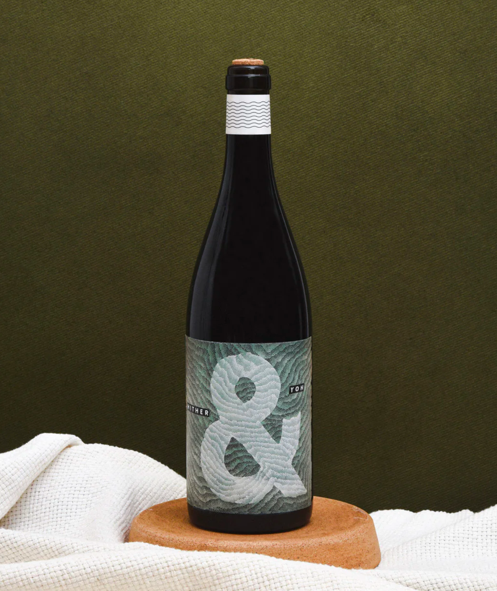 A bottle of Hither & Yon Syrah sitting on a wood base and woven white rug with a green background.