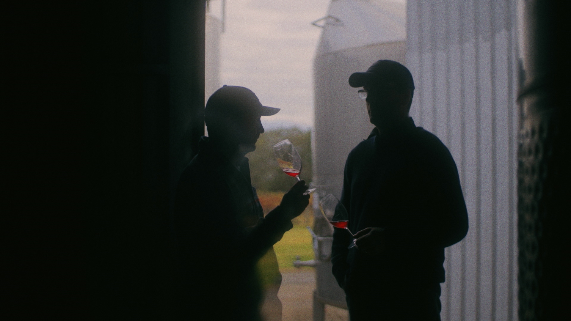 Richard and Malcolm Leask standing in a winery shed with a glass of red wine in hand.