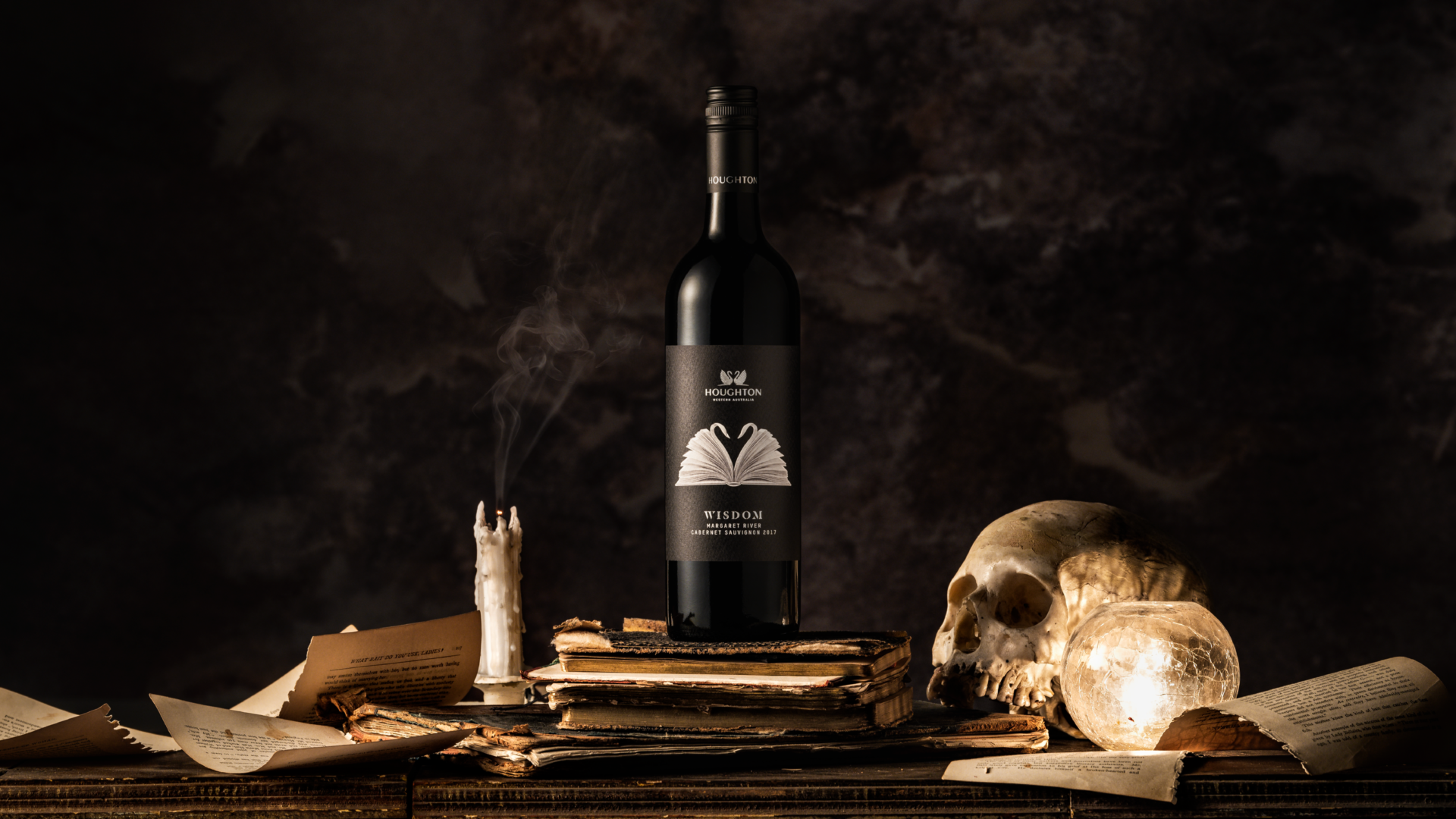 A bottle of Houghton Wisdom wine sits on top of an old book with torn pages all around, a skull and a tall candle. 