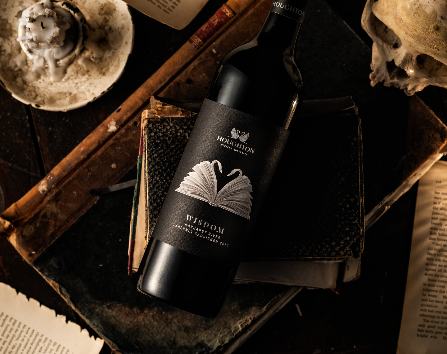 A close up of the Houghton Wisdom wine bottle featuring packaging design featuring an open book illustration with pages folded to mimic the Houghton double swan logo and elements of silver foil on a black background.