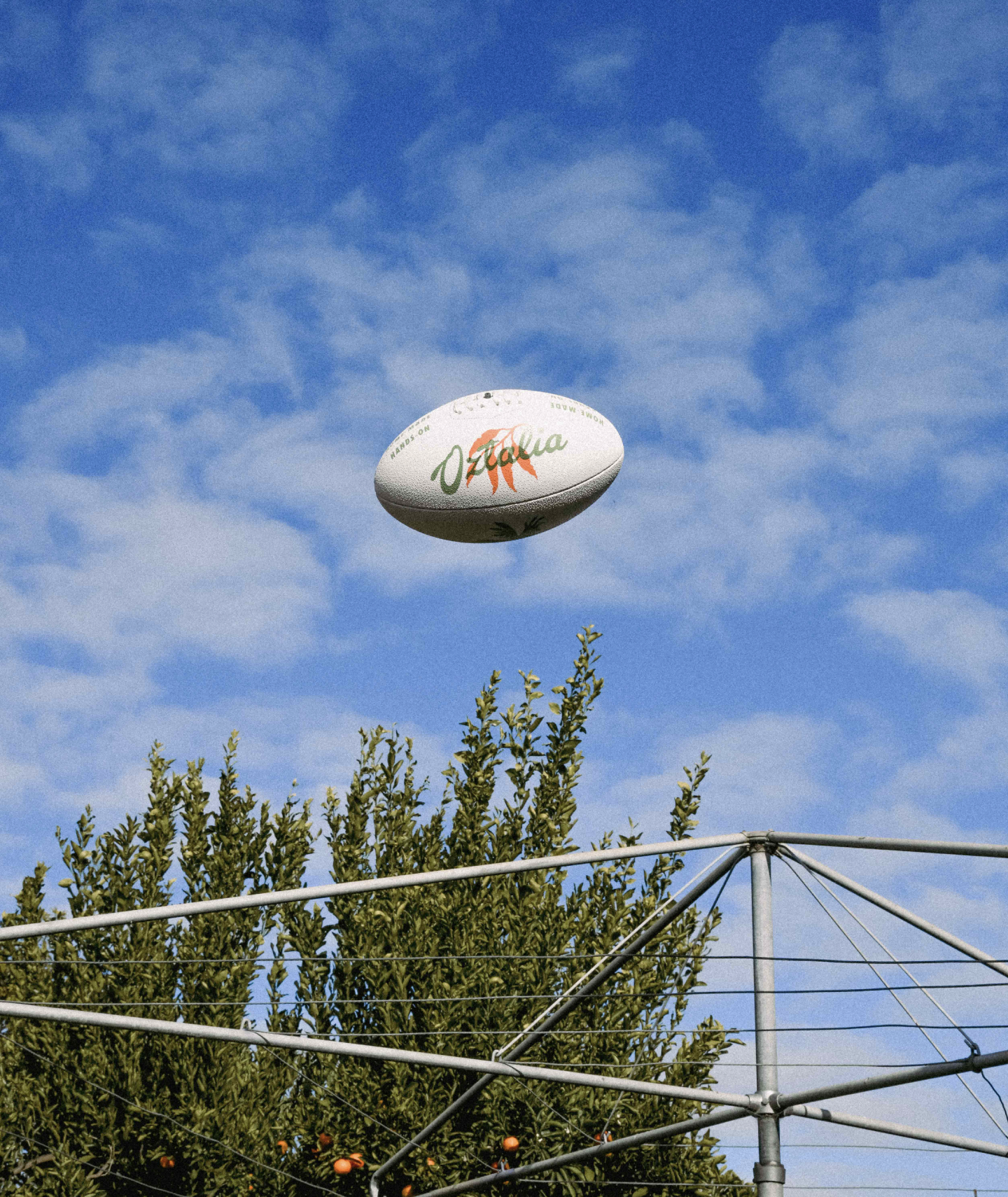 A photograph of an Oztalia branded football in the blue sky over a hills hoist clothesline with an orange tree in the background. 