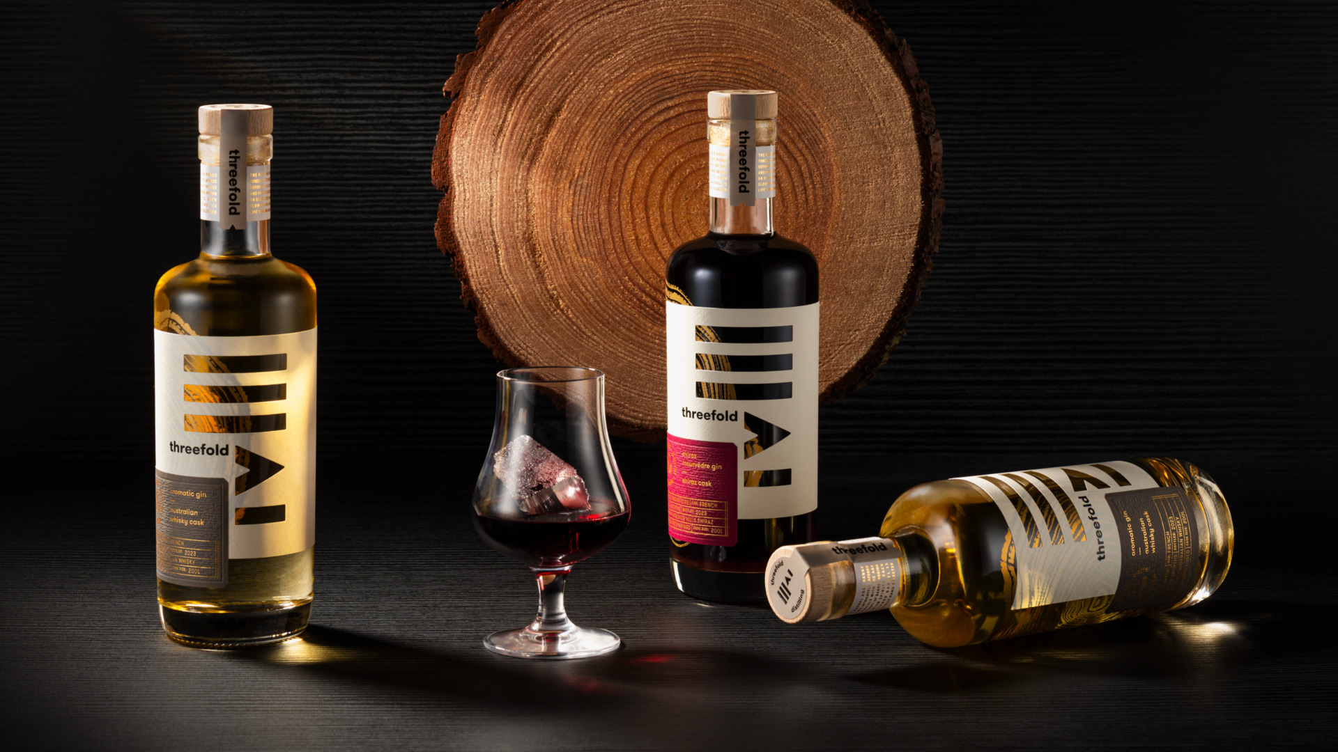 3 Threefold Barrel Aged gin bottles on a black backdrop showcasing a diecut label with gold foil over a screen-printed wood pattern. There is a woodcut in the background and a glass of gin over ice. 