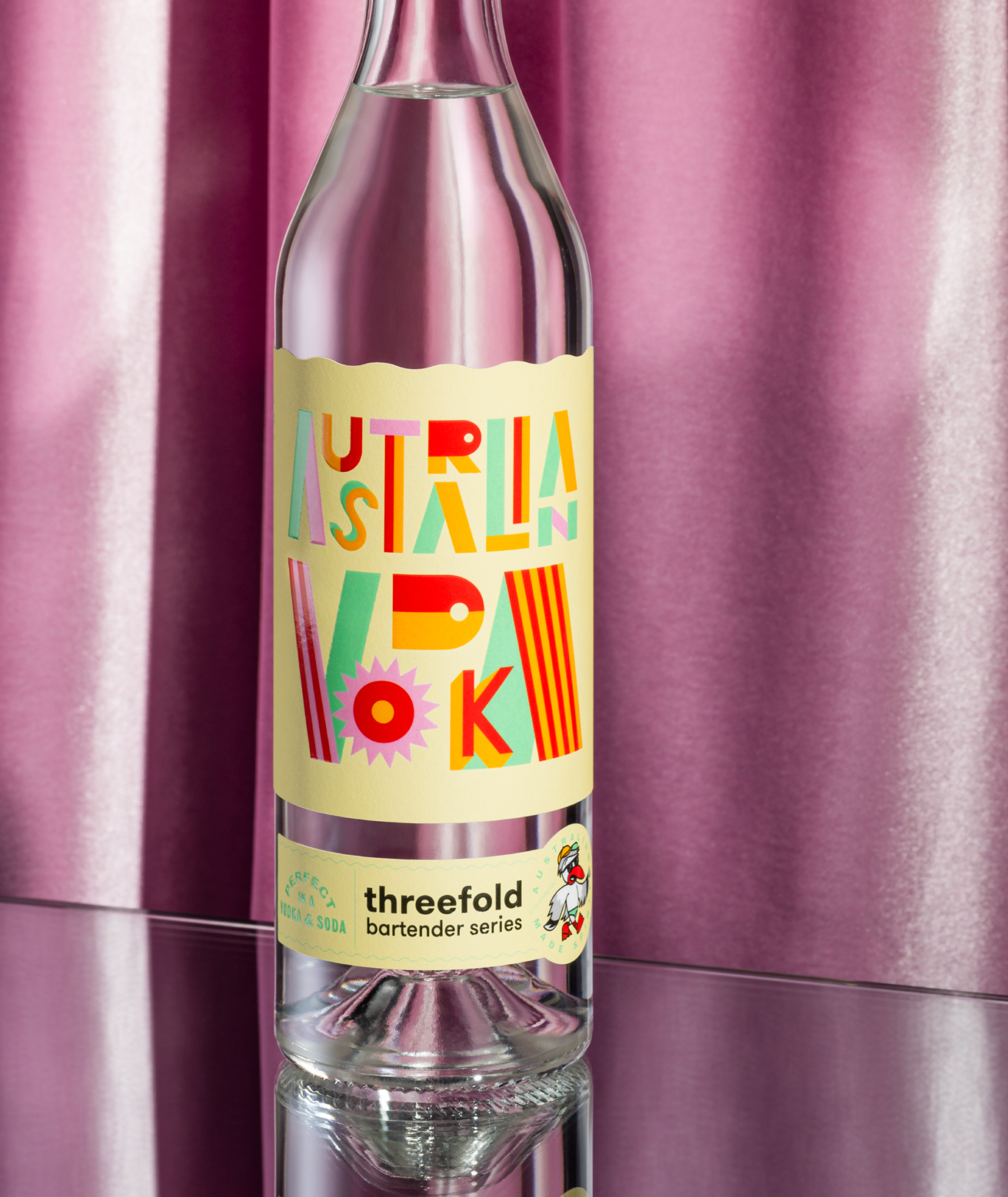 A bottle of Threefold Bartender's Series Australian Vodka with a light yellow label on a mirrored base with a pink fabric background.