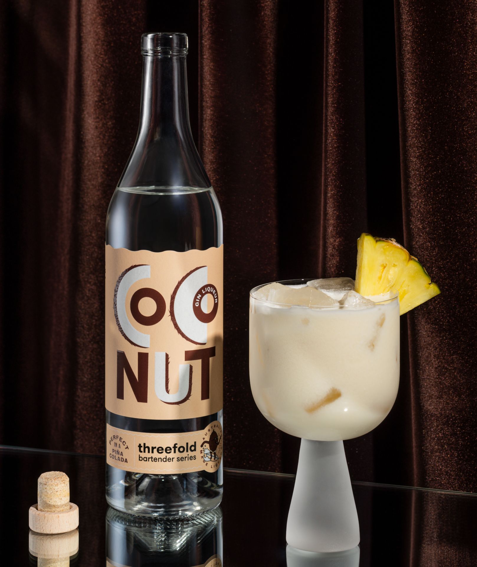 An open bottle of Threefold Bartender's Series Coconut Gin with a beige label on a mirrored base with a brown fabric background next to a Piña Colada over ice with a pineapple garnish.
