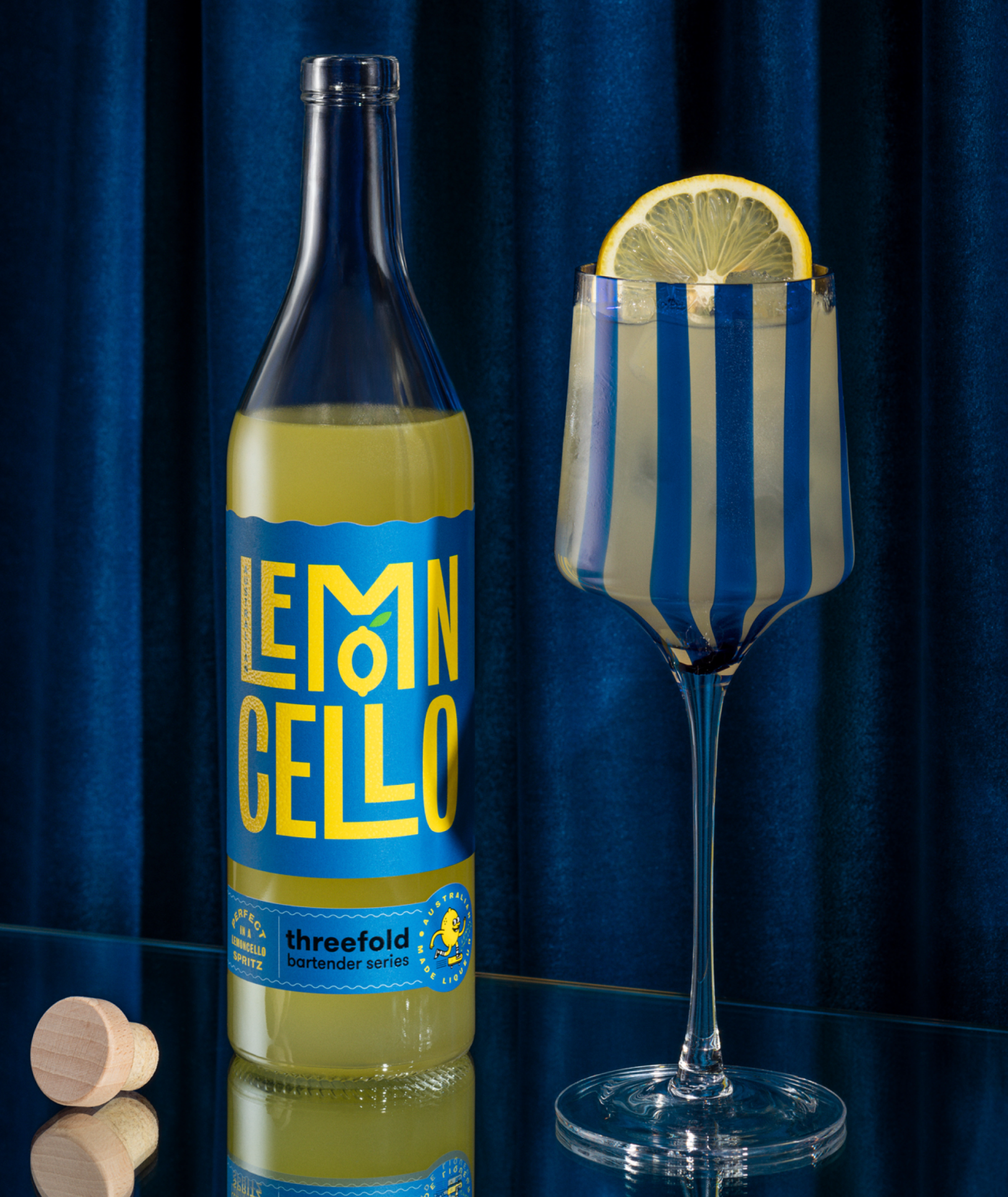 An open bottle of Threefold Bartender's Series Lemoncello with a blue label on a mirrored base with a navy fabric background next to a striped tall glass of Lemoncello over ice with a lemon garnish. 