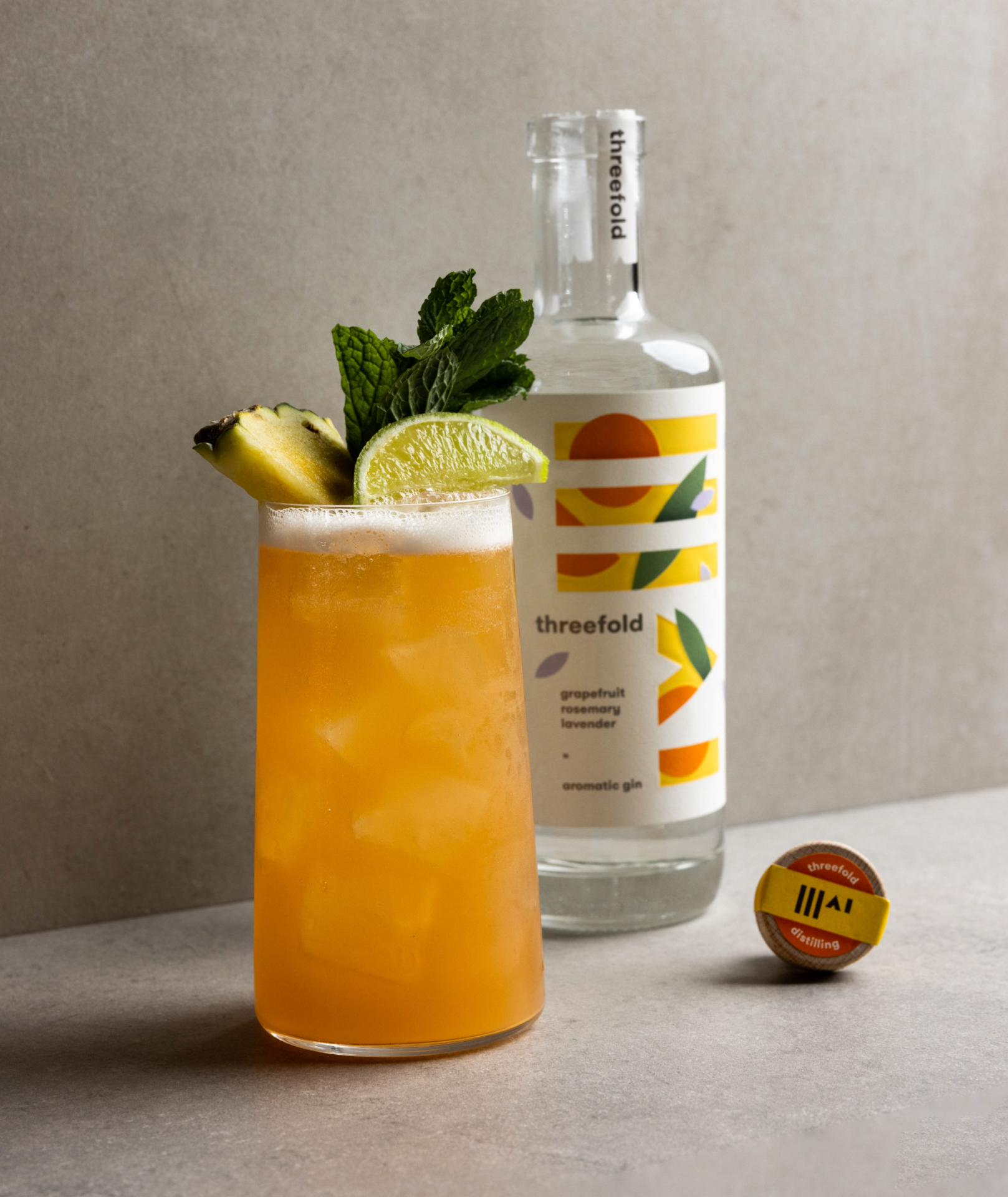 An open bottle of Threefold Aromatic gin next to a cocktail in a tall glass with a mint, lime and pineapple garnish.