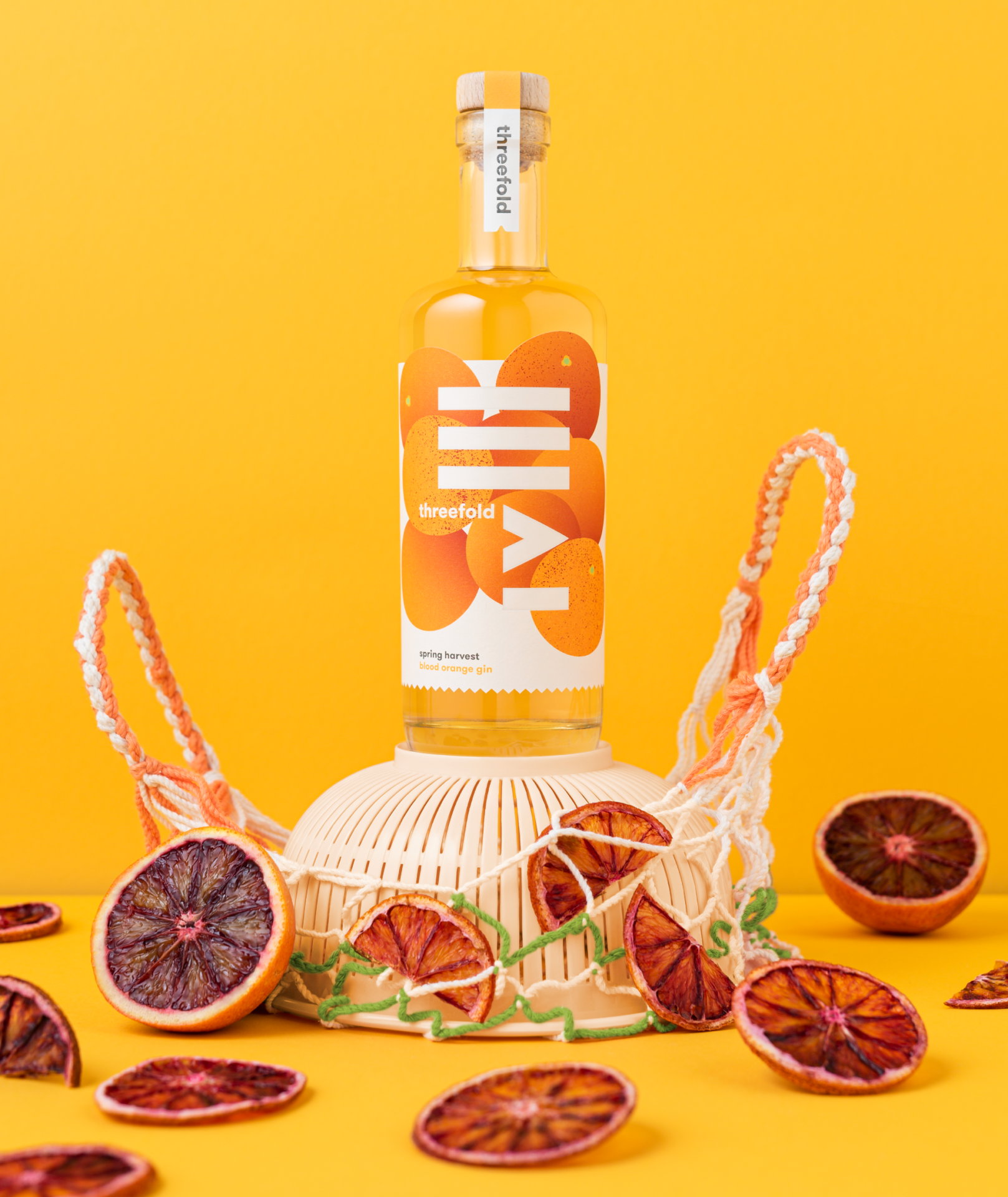A photograph of Threefold Spring Harvest Blood Orange gin with bright packaging design, perched on a plastic sieve and rope a shopping bag with an abundance of blood oranges scattered around.