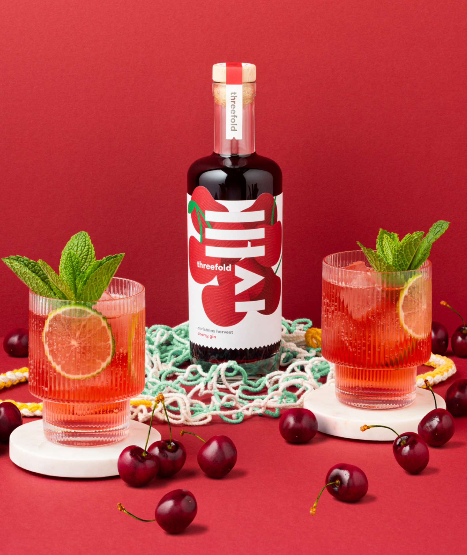 A photograph of Threefold Christmas Harvest Cherry gin with bright packaging design, perched on a plastic sieve and rope a shopping bag with two cocktails featuring mint and lime garnish and an abundance of cherries scattered around.