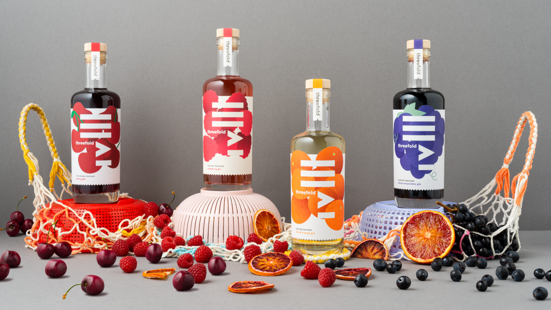 A photograph of 4 bottles of Threefold Harvest Series gin with bright packaging design, perched on plastic sieves and rope shopping bags with an abundance of fruit scattered around. 