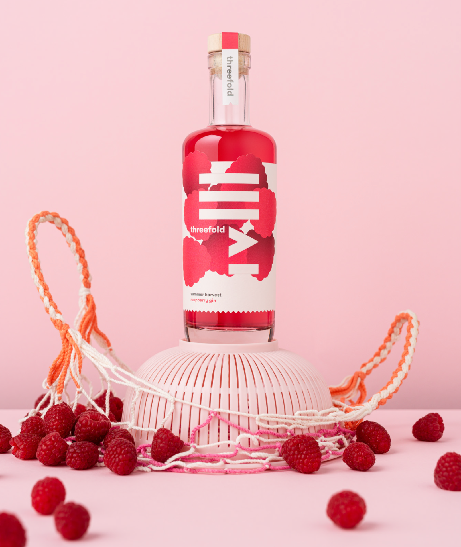 A photograph of Threefold Summer Harvest Raspberry gin with bright packaging design, perched on a plastic sieve and rope a shopping bag with an abundance of raspberries scattered around.