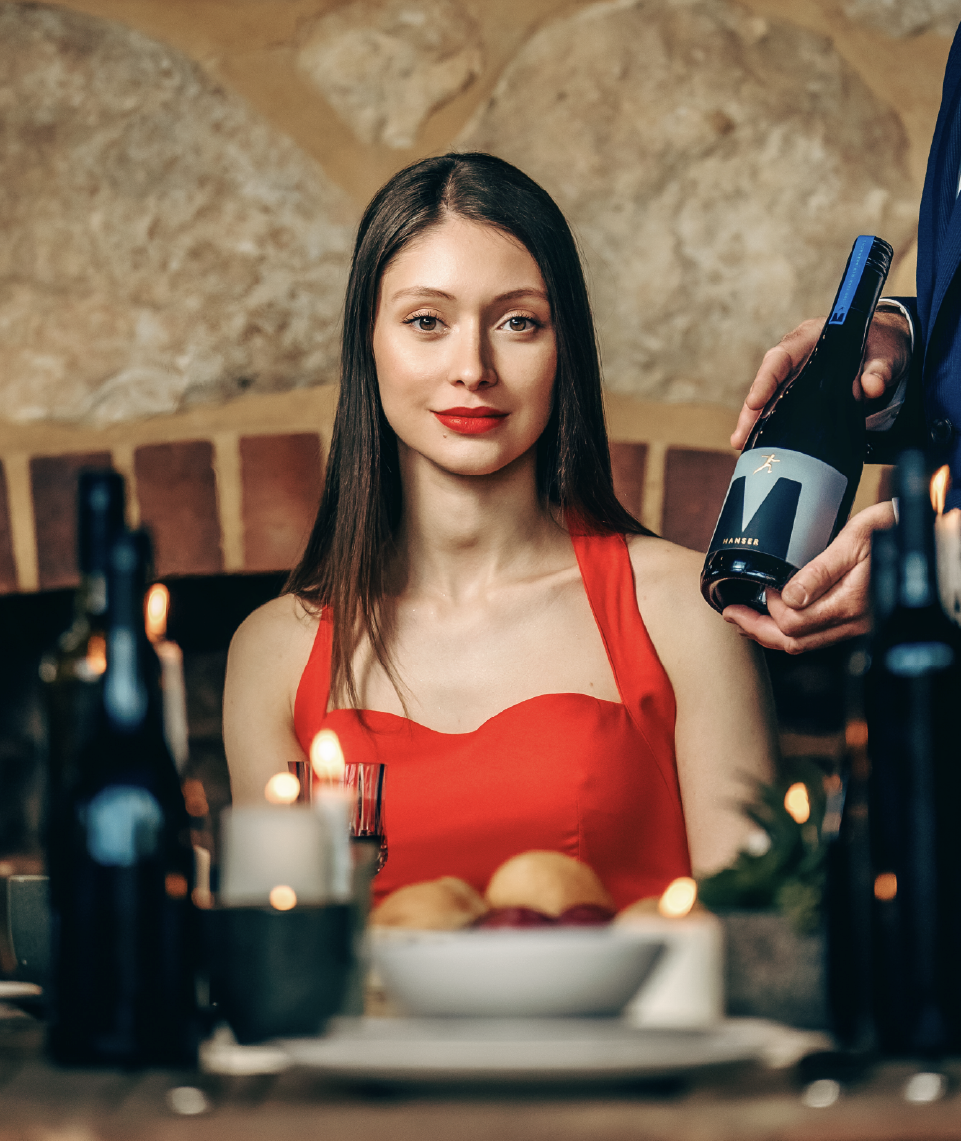 Model seated at a table in a bright red dress being presented with a bottle of Manser Wines, showcasing a premium wine brand and lifestyle setting.
