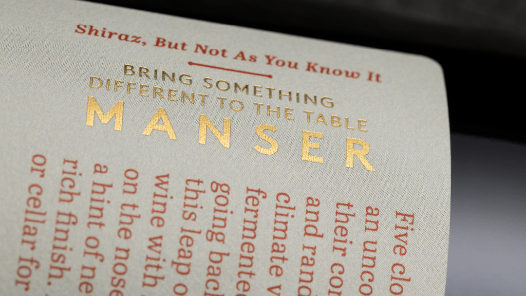 Close-up detail of gold foil on a premium wine label, highlighting brand positioning copy: “Bring something different to the table” for Manser Wines.