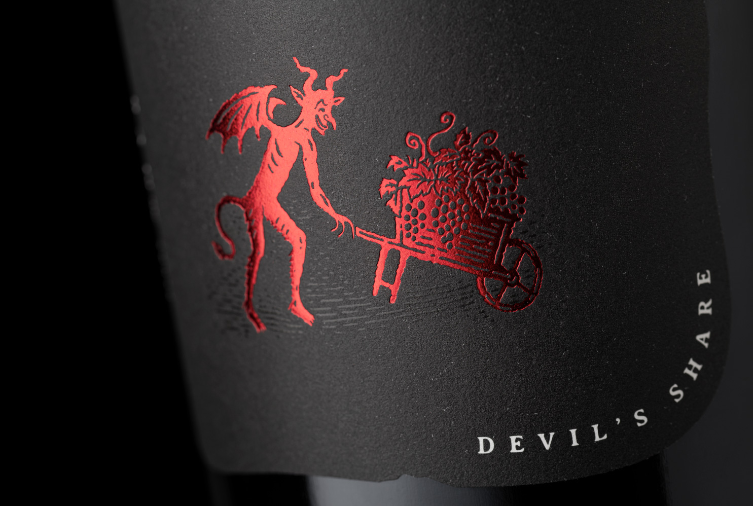 Extreme close-up of the 'Devil's Share' icon for The Devil Made Pinot Single Clone, featuring precise red metallic foil stamping of a devil pushing a wheelbarrow on a textured matte black label.