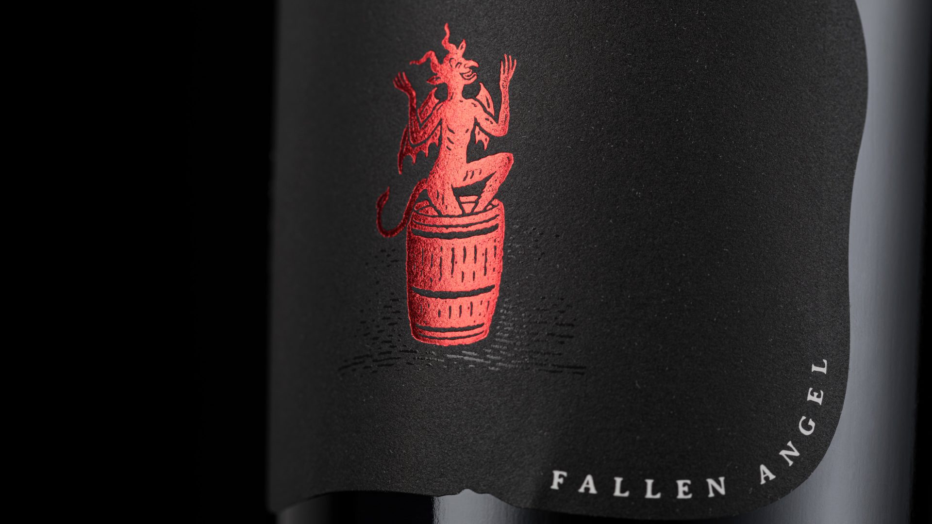 Extreme close-up of the 'Fallen Angel' brand icon for The Devil Made Pinot Single Clone, featuring vibrant red metallic foil stamping on a textured matte black label.