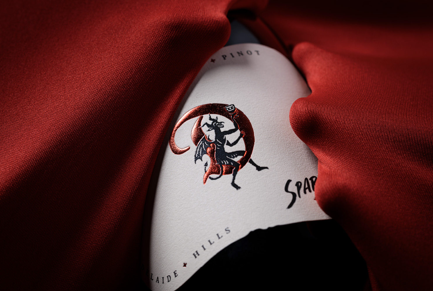 Close-up of the custom brand icon for The Devil Made Pinot Sparkling, featuring a devil character perched on a red metallic foil.