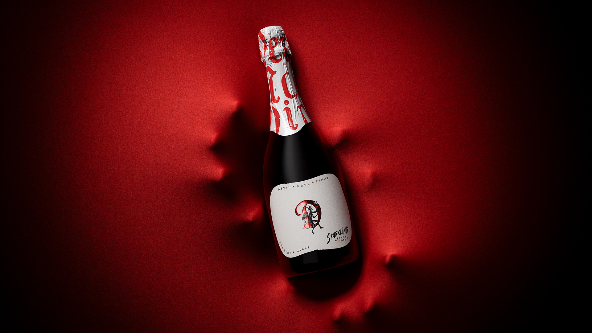 Custom sparkling wine label design for The Devil Made Pinot, featuring a devil illustration on an Adelaide Hills Sparkling Pinot Noir bottle against a soft red background
